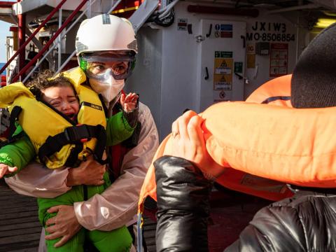 La sage-femme de l'équipage tient dans ses bras un jeune enfant qui pleure tandis que la mère de celui-ci enlève son gilet de sauvetage. Ils viennent d'être secourus d'une embarcation pneumatique en péril contenant plus de cent personnes.