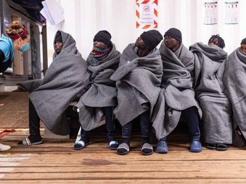 Des rescapés enroulés dans des couvertures sont assis sur un banc et attendent leur tour pour une consultation médicale dans la clinique du bateau. L'un d'entre eux discute avec la sage-femme de l'équipage.